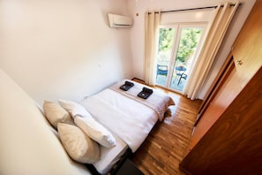 Ein Schlafzimmer mit Bett, Kissen, Nachttisch, Klimaanlage und Blick ins Freie durch eine Glastür.