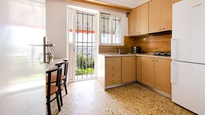 A kitchen with wooden cabinets, a white refrigerator, a dining table with chairs, and a window with metal bars. - Acropolis Hillside Escape, Sea & Nature Views (Athina)