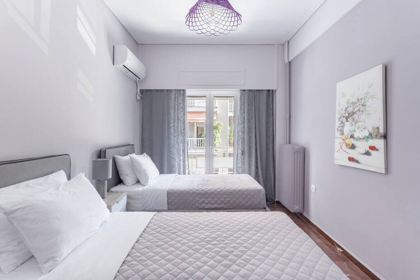 A hotel room with two beds, a window with curtains, a radiator, and a wall-mounted air conditioner.