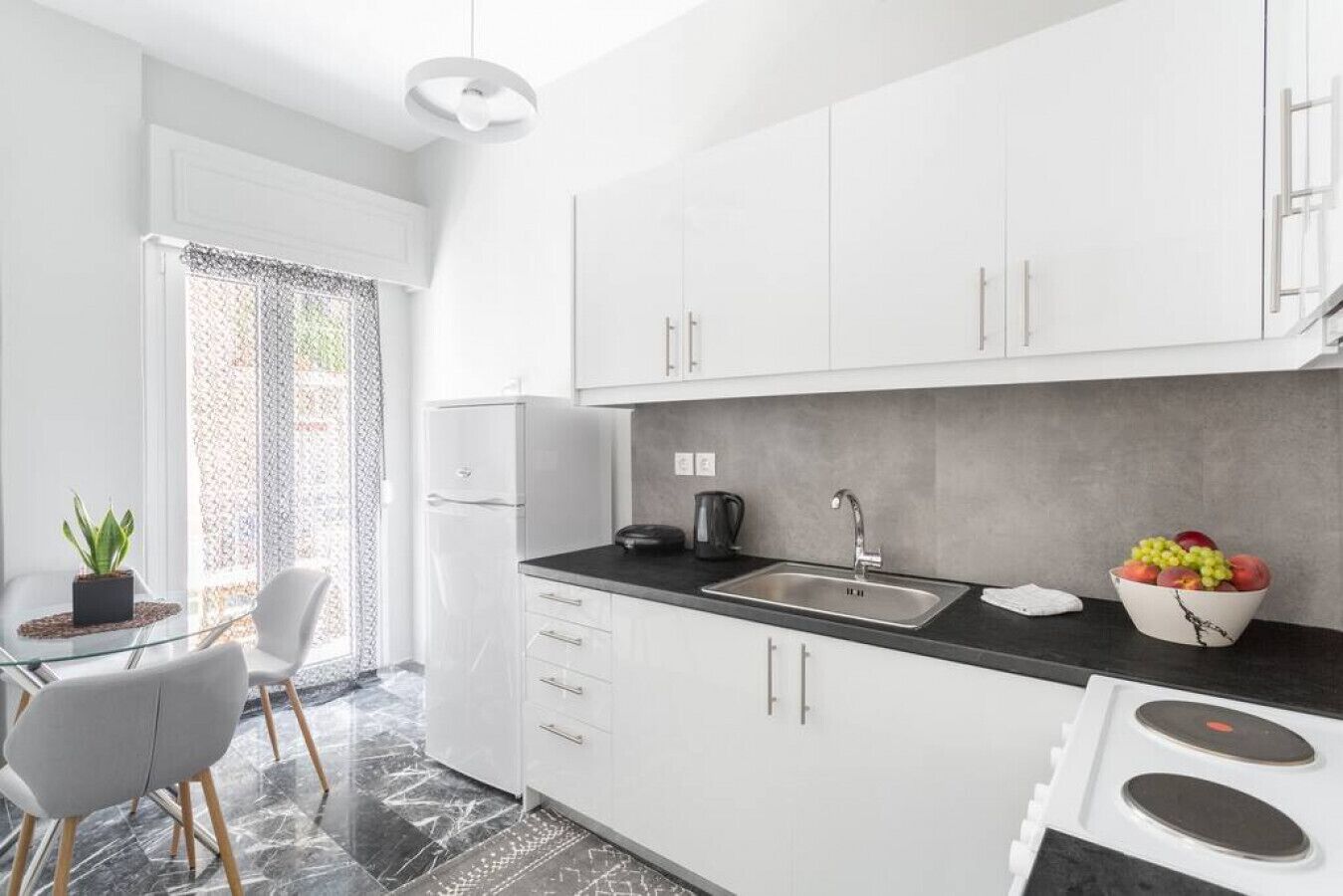 Una cocina moderna con armarios blancos, encimera negra, un lavabo y una estufa. Hay una planta en maceta a la izquierda y un cuenco con fruta a la derecha.