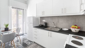 A modern kitchen with white cabinets, a black countertop, a sink, and a stove. There is a potted plant on the left and a bowl of fruit on the right. - Explore History from a Chic Central Apartment (Athina)