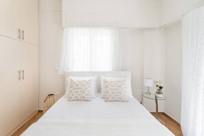 A minimalist bedroom with a large bed, white bedding, and a small bedside table with a lamp and flowers. - Explore History from a Chic Central Apartment (Athina)