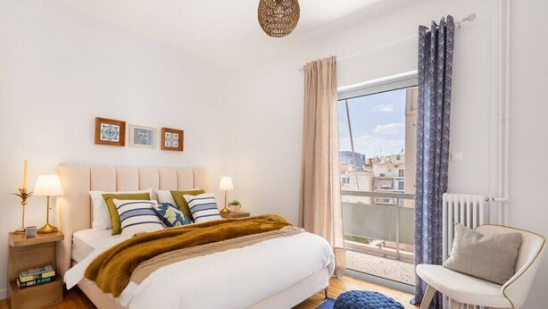 Een slaapkamer met een bed, nachtkastjes, een stoel, een raam met gordijnen en uitzicht op gebouwen.