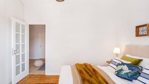 Een slaapkamer met een bed, een nachtkastje, een lamp, een aan de muur hangend schilderij en een deur die naar een andere kamer leidt.