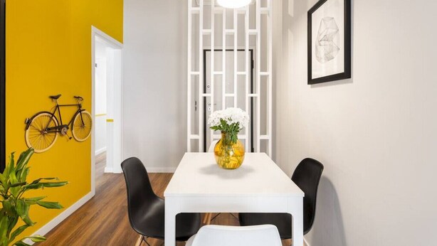 Un espace repas avec une table blanche, des chaises noires, un mur d’accent jaune, un vélo et une suspension blanche.