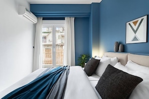Ett hotellrum med en säng, ett fönster med gardiner, en väggmonterad luftkonditioneringsanläggning och en inramad tavla på väggen.