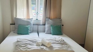 A neatly made twin bed with white bedding and two pillows, one teal and one white, in a room with a window and curtains.