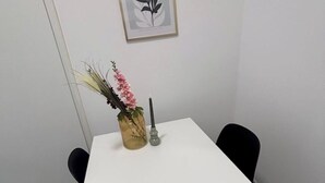 A white table with a vase containing pink flowers and a candle, and a framed plant on the wall.