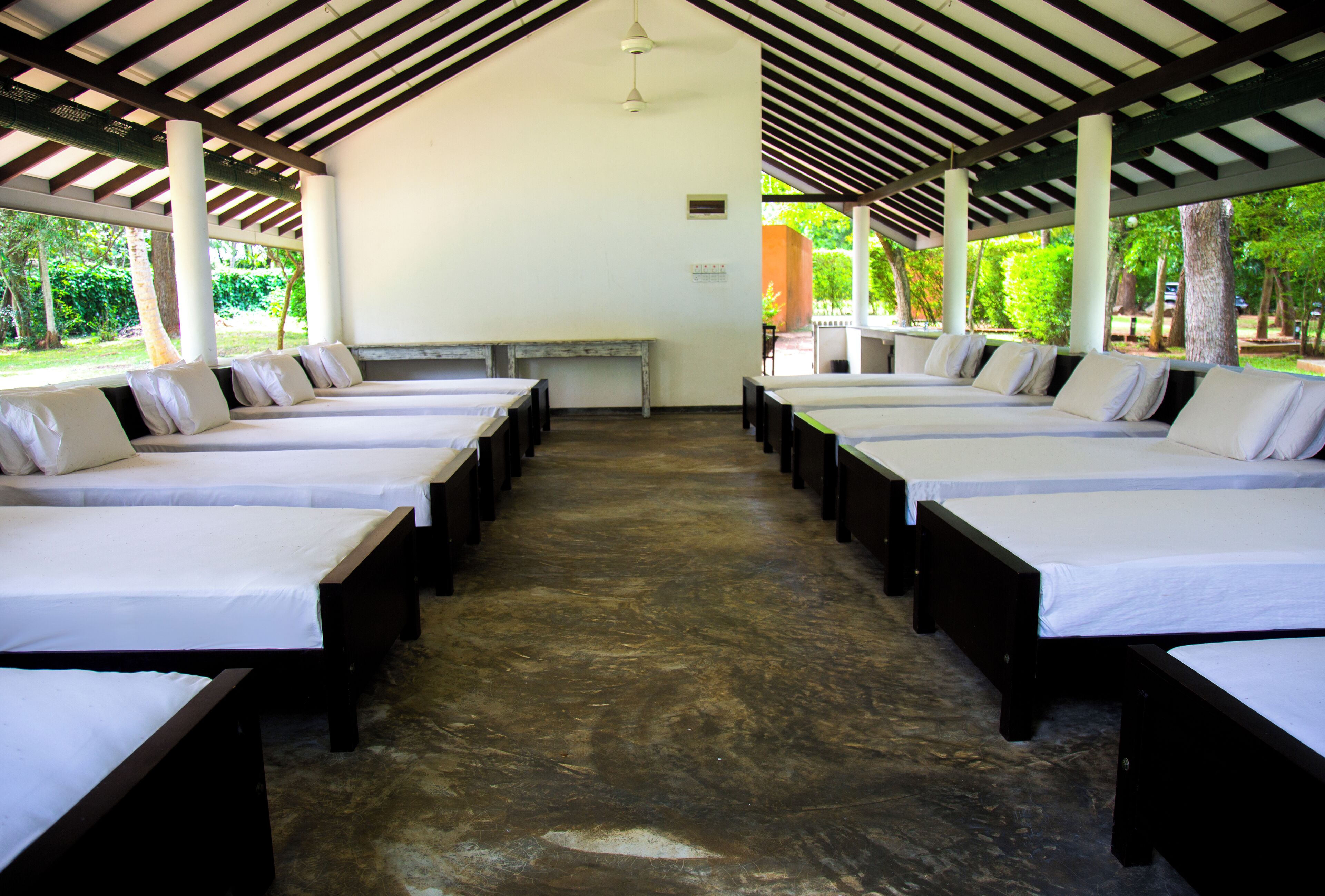 A row of single beds with white linens, wooden headboards, and a concrete floor under a striped roof structure.