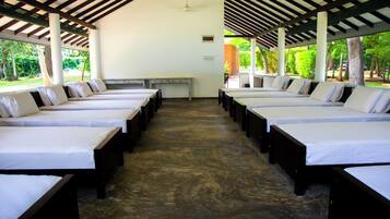 A row of single beds with white linens, wooden headboards, and a concrete floor under a striped roof structure.