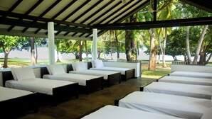 Area relax esterna coperta con diversi lettini disposti a file, affacciata su uno specchio d'acqua e circondata da alberi.
