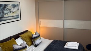 Cama bem arrumada com colcha escura, travesseiros brancos e uma toalha dobrada ao lado.
