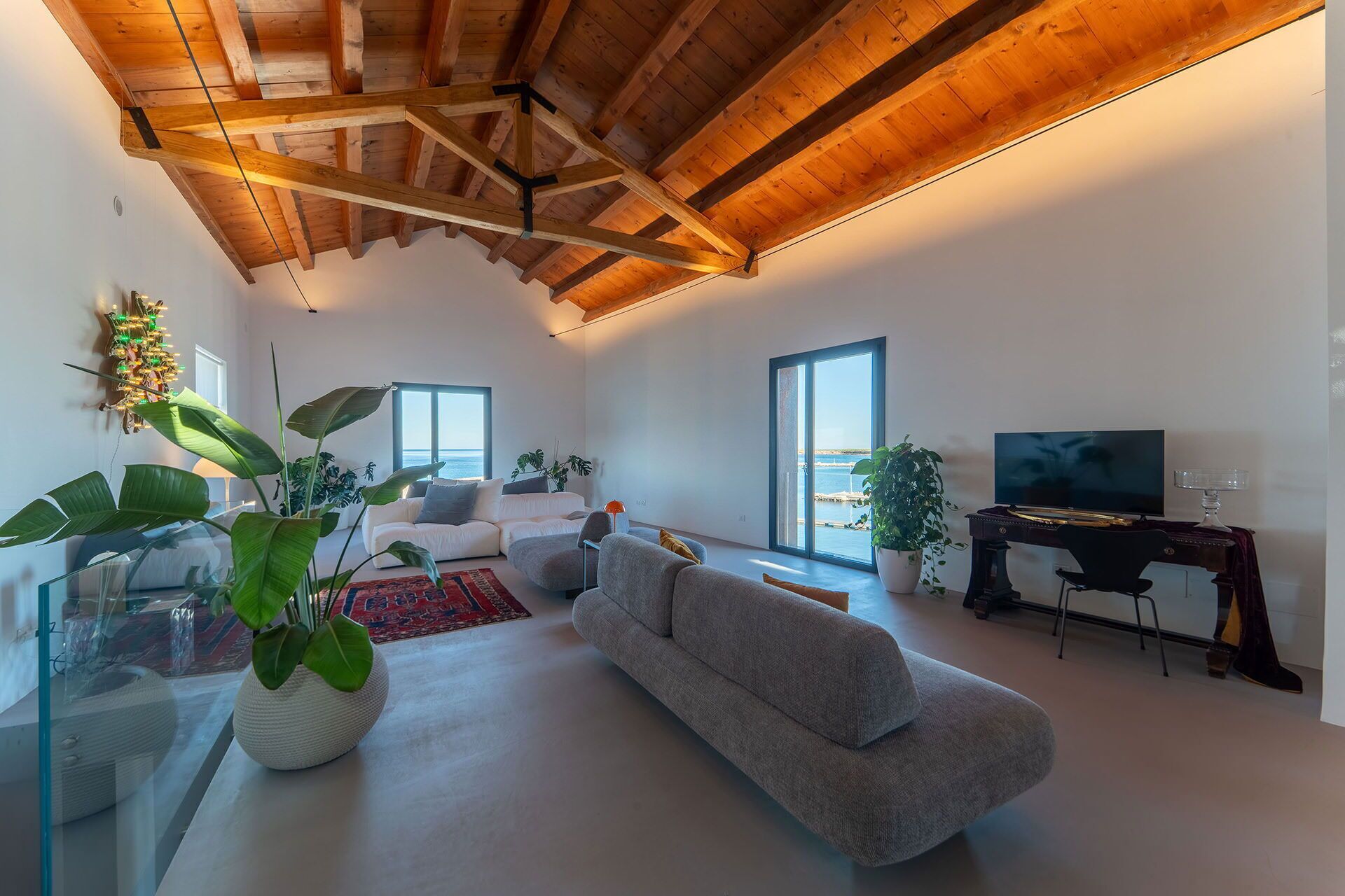 Ein geräumiges Wohnzimmer mit hoher Decke, einem großen Fenster mit Meerblick, einer bequemen Couch, einem Fernseher und einem Schreibtisch mit Computer.