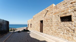 Ein Steingebäude mit Blick auf das Meer, ein gepflasterter Weg und ein Metallzaun.