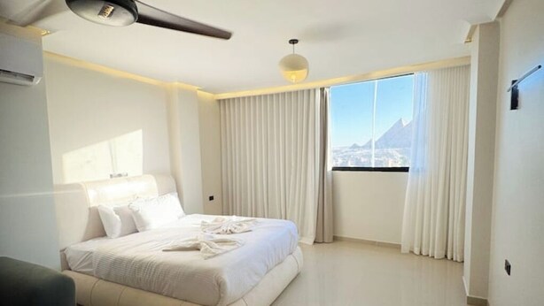 A modern hotel room with a large bed, a ceiling fan, a window with curtains, and a view of a mountain. - Modern Deluxe Studio by the Pyramids – Comfort & Style in Giza (Giza Governorate)