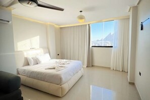 A modern hotel room with a large bed, a ceiling fan, a window with curtains, and a view of a mountain. - Modern Deluxe Studio by the Pyramids – Comfort & Style in Giza (Giza Governorate)