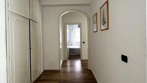 Een gang met houten vloer, witte muren en een gewelfde deuropening naar een andere kamer.