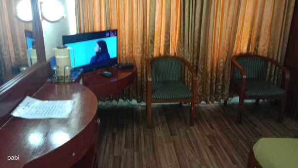 A hotel room with a wooden desk, a television, two green upholstered chairs, and a large window with curtains. - Hotel Goutham Manor (Chennai)