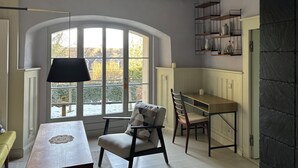 Um quarto com uma grande janela em arco, uma mesa de jantar com cadeiras, um abajur e uma estante com objetos decorativos.