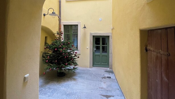 Uma viela estreita com um pinheiro de Natal decorado com enfeites vermelhos, uma porta verde e um edifício amarelo com uma janela e um candeeiro.