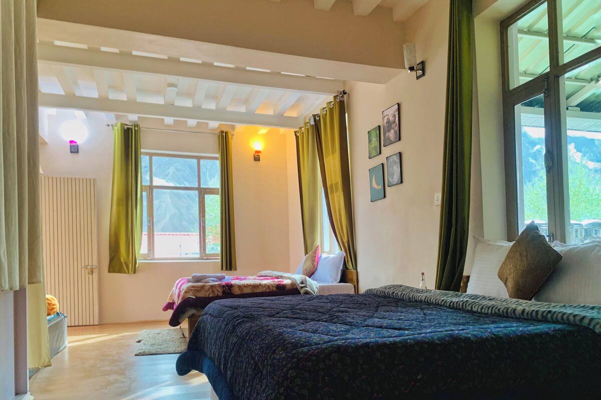 A bedroom with a large bed, a window with curtains, a door, and a wall-mounted light.