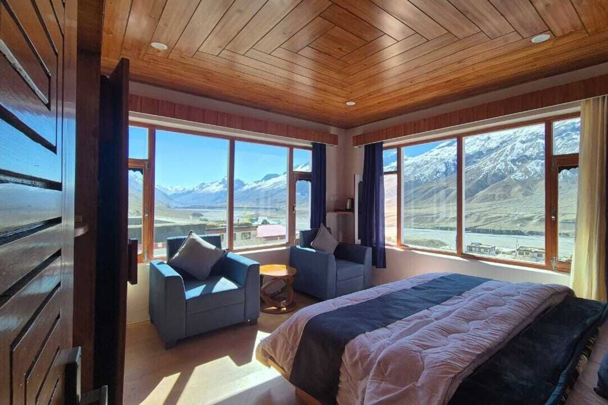 A bedroom with a large bed, two armchairs, a small table, and a view of mountains through large windows.