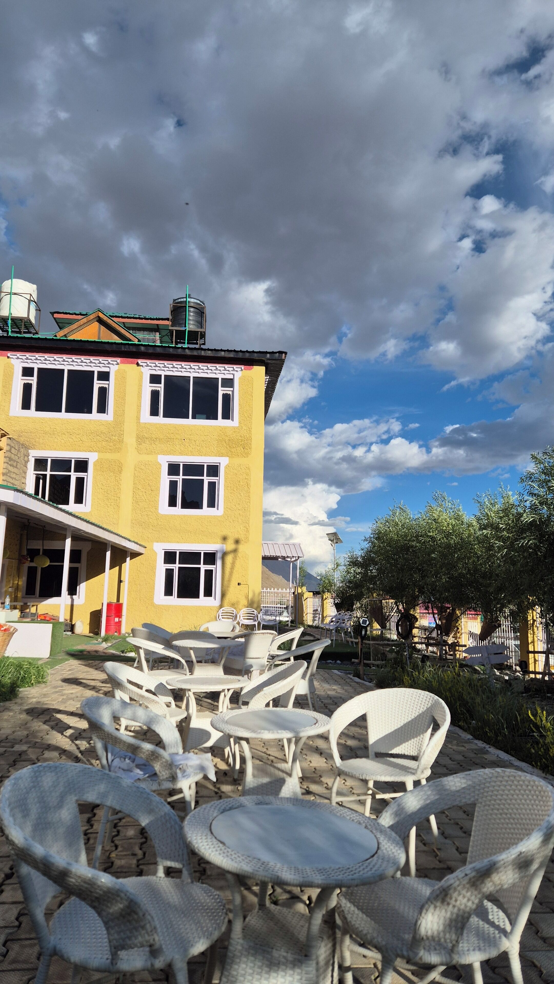 A yellow building with multiple windows and a green roof, outdoor seating with white wicker chairs and tables, and a clear sky with clouds.
