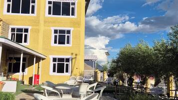 A yellow building with multiple windows and a green roof, outdoor seating with white wicker chairs and tables, and a clear sky with clouds.