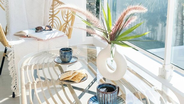 Un tavolo rotondo in vetro con bordi in rattan intrecciato, due tazze di ceramica, un vaso con una pianta e un libro con occhiali da sole su una sedia in vimini.