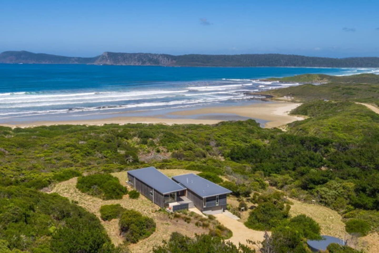 A modern two-story house with a flat roof, surrounded by dense green vegetation and a beach in the background.