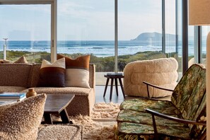 A modern living room with a large window overlooking a coastal view, featuring a sofa, armchair, and a small table. - Cloudy Bay Beach House – Luxury Escape in Bruny (South Bruny)