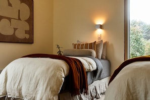 A bedroom with two beds, a bedside table with a lamp, and a window with a view of greenery.