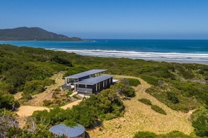 A modern house with a flat roof, surrounded by dense vegetation and a beach. - Cloudy Bay Beach House – Luxury Escape in Bruny (South Bruny)