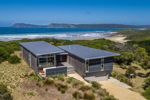A modern two-story house with a large glass window on the ground floor, situated on a sandy terrain with a view of the ocean and hills in the background.