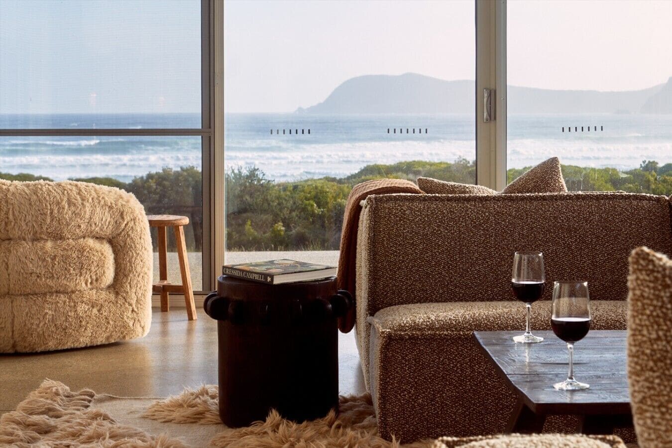 A cozy seating area with a view of the ocean, a book on a stool, and two glasses of red wine on a table.