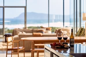 A modern living room with a large window overlooking a scenic view, a sofa, a dining table, and two glasses of red wine.