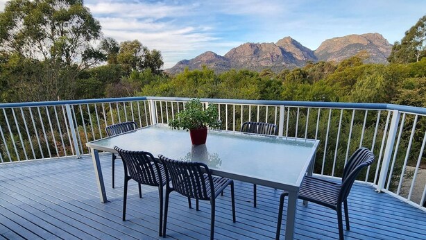 A deck with a glass-top table and chairs, surrounded by a white railing, overlooking a mountainous landscape. - Forest Edge - Breathtaking Views of the Hazards (Coles Bay)