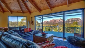 A spacious living room with a leather sofa, a coffee table, and a view of mountains through large windows. - Forest Edge - Breathtaking Views of the Hazards (Coles Bay)