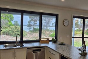 A kitchen with a view of a lake, a clock on the wall, and a dishwasher. - Joeys Place- Waterfront| Views| Sunsets @Freycinet (Coles Bay)