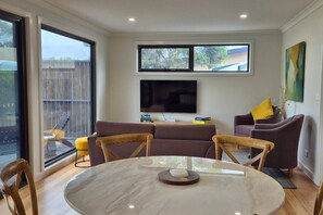 A modern living room with a large TV, two sofas, a dining table, and a balcony with a view. - Joeys Place- Waterfront| Views| Sunsets @Freycinet (Coles Bay)