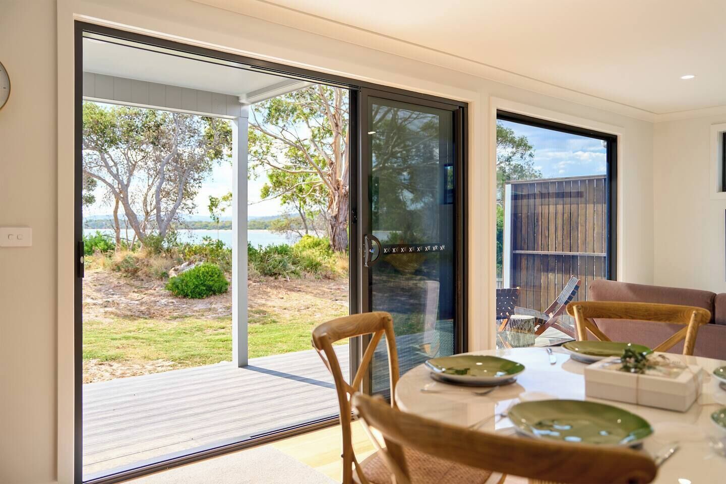 A dining area with a table set for two, large sliding glass doors leading to an outdoor deck with a view of trees and a body of water.