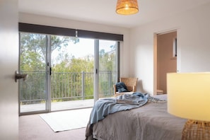 Une chambre avec une grande porte-fenêtre coulissante donnant sur un balcon, un lit avec une couverture, une chaise en osier et une lampe.