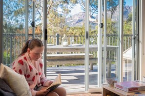 Une personne assise à l’intérieur, avec un livre, une boîte de jeu de Monopoly, et une vue sur une terrasse et le paysage extérieur.