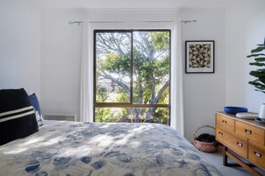 Une chambre avec une grande fenêtre, un lit recouvert d’une couvre-lit à motifs, une commode et un tableau encadré accroché au mur.