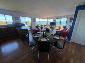 A dining area with a round table set for six, a kitchen area, and large windows offering a view of the sea. - Aperture, Stunning Views Frame Your Escape (Coles Bay)