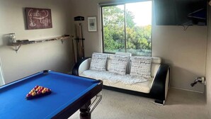 Ein Zimmer mit Billardtisch, einer Couch mit Zierkissen, einem Fenster mit Blick ins Grüne und einem an der Wand befestigten Fernseher.