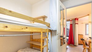 Une chambre avec un lit superposé en bois, un matelas blanc et une porte donnant sur un coin repas avec une table et des chaises.