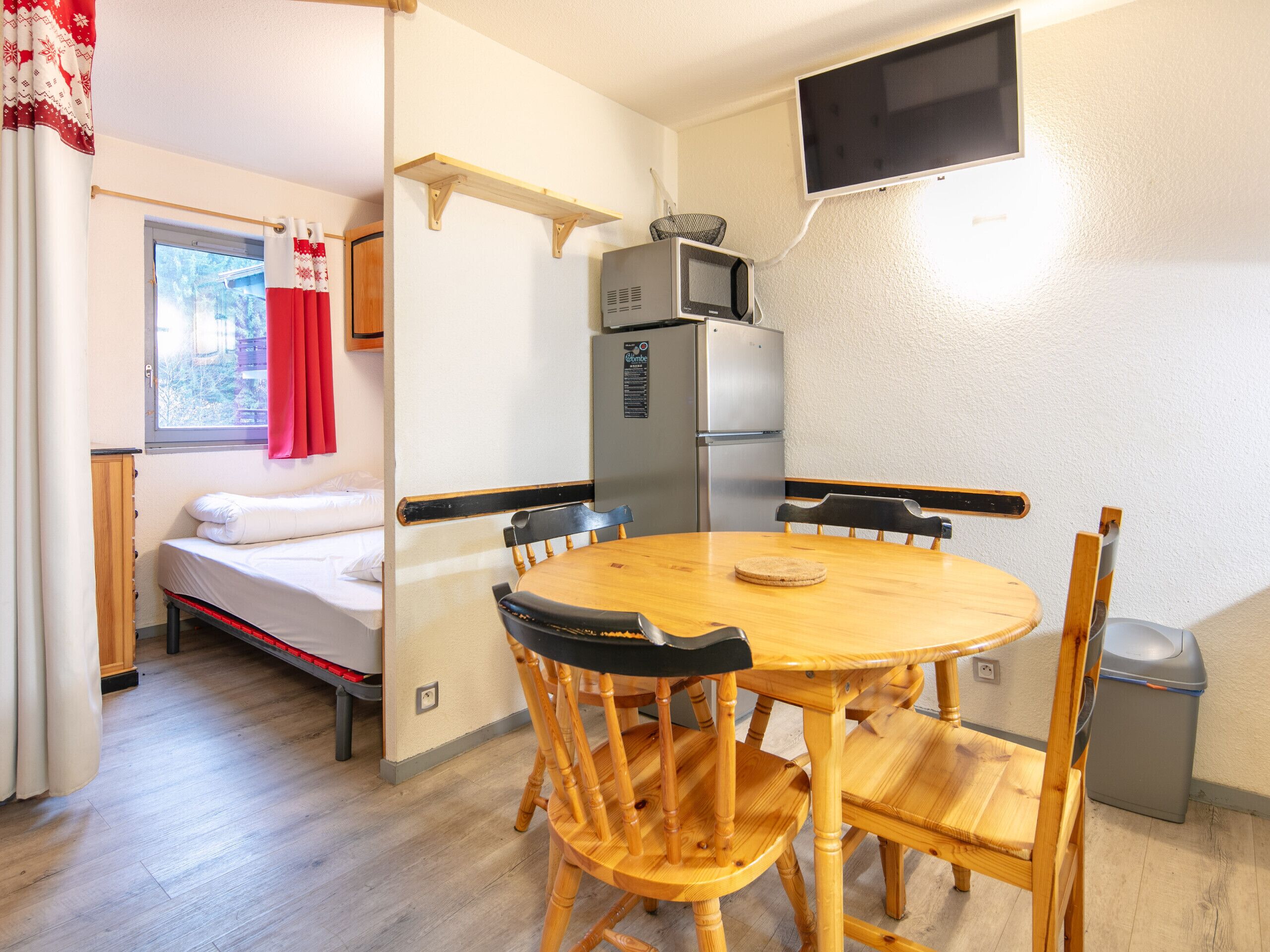 A compact room with a bed, a dining table with chairs, a microwave, a refrigerator, a TV mounted on the wall, and a window with curtains.
