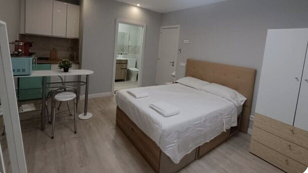 Un dormitorio moderno con una cama individual, una mesita de noche, un escritorio pequeño con una silla y una kitchenette al fondo.