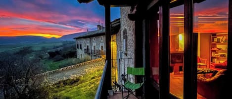 A balcony with a view of a sunset over a rural landscape, a stone building, and a room with a large window.